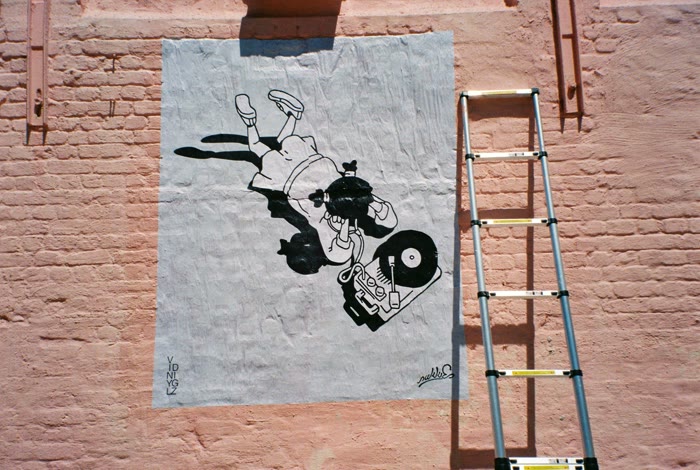 Fresh wheatpaste poster going up beside the ladder, mid-install
